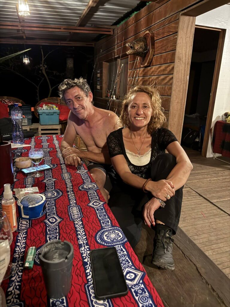 A man and a woman are sitting at a table covered with a red and blue patterned cloth, with fishing rods on the wall behind them.