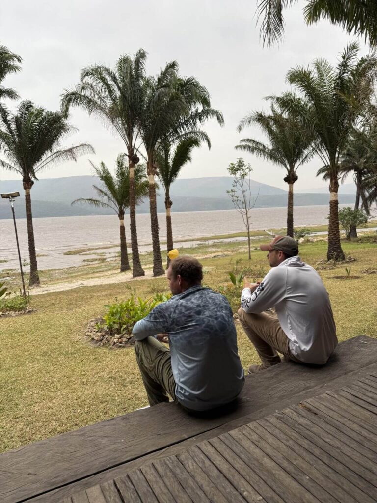 Two men sit on a wooden deck overlooking a lake with palm trees under a cloudy sky.