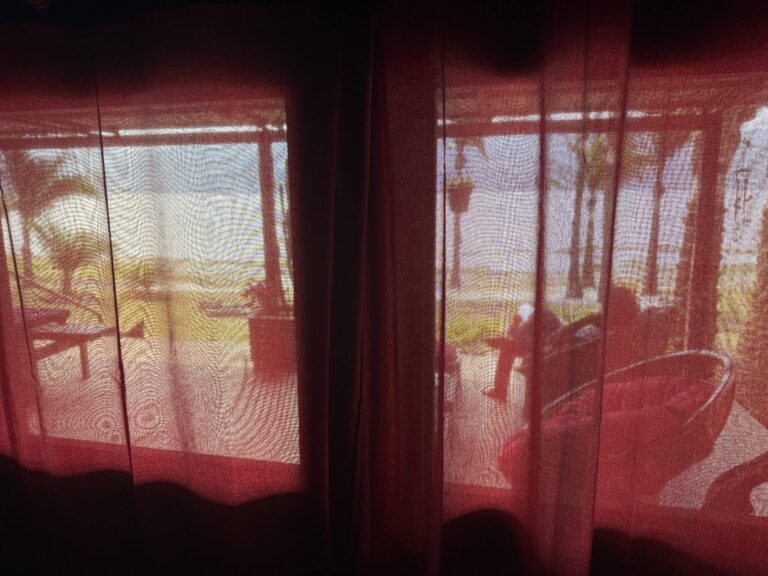 Red sheer curtains partially obscure a sunny outdoor patio scene with a person sitting in a chair.