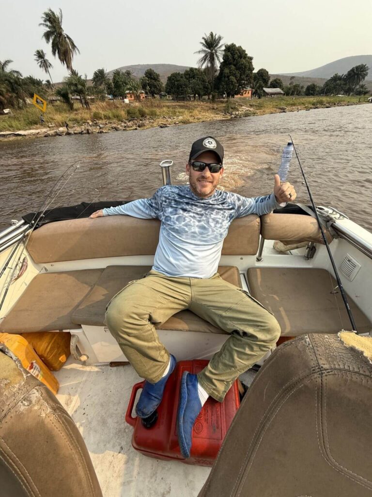 A man wearing sunglasses and a baseball cap gives a thumbs-up while sitting on a boat with a red gas can between his legs.