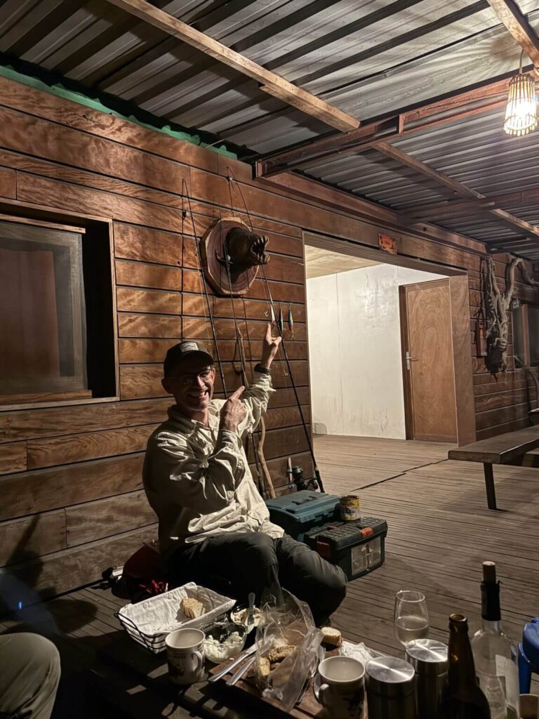 A man sits on a wooden deck, pointing at fishing lures displayed on the wall next to a mounted bear head.