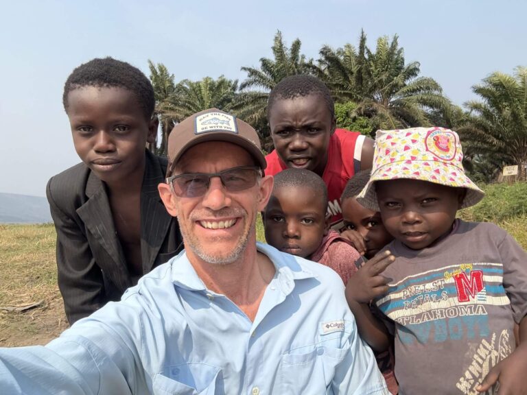 A smiling man takes a selfie with a group of children in front of palm trees.