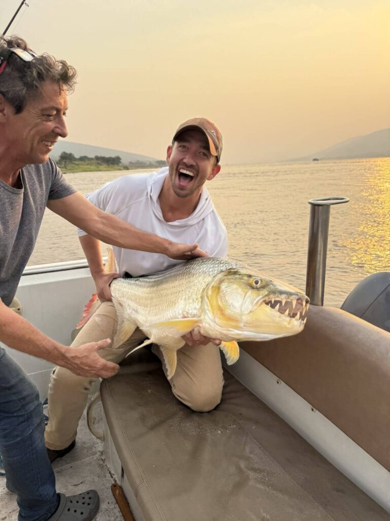 Two men excitedly show off a large, predatory fish with sharp teeth on a boat at sunset.