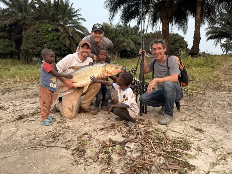 Three adults and two children pose with a large fish and fishing rods near palm trees.