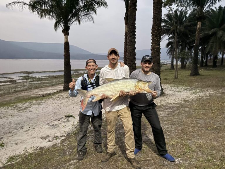Three men proudly display a large fish they caught by a lake surrounded by palm trees.