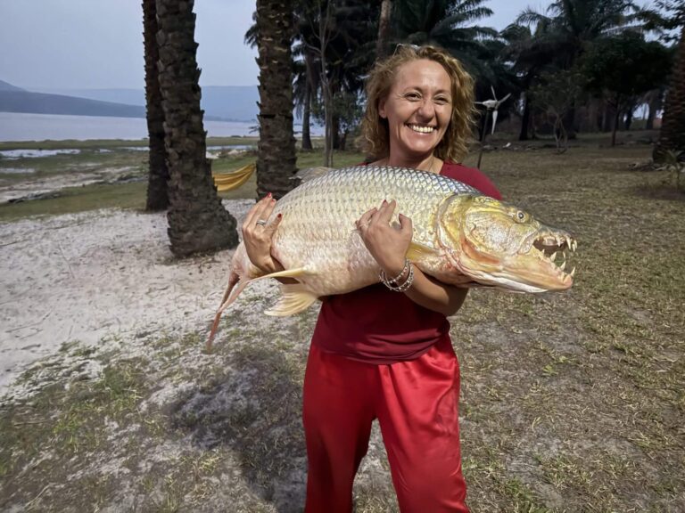 A woman proudly holds a large, formidable-looking fish with sharp teeth in a tropical setting with palm trees and a lake.