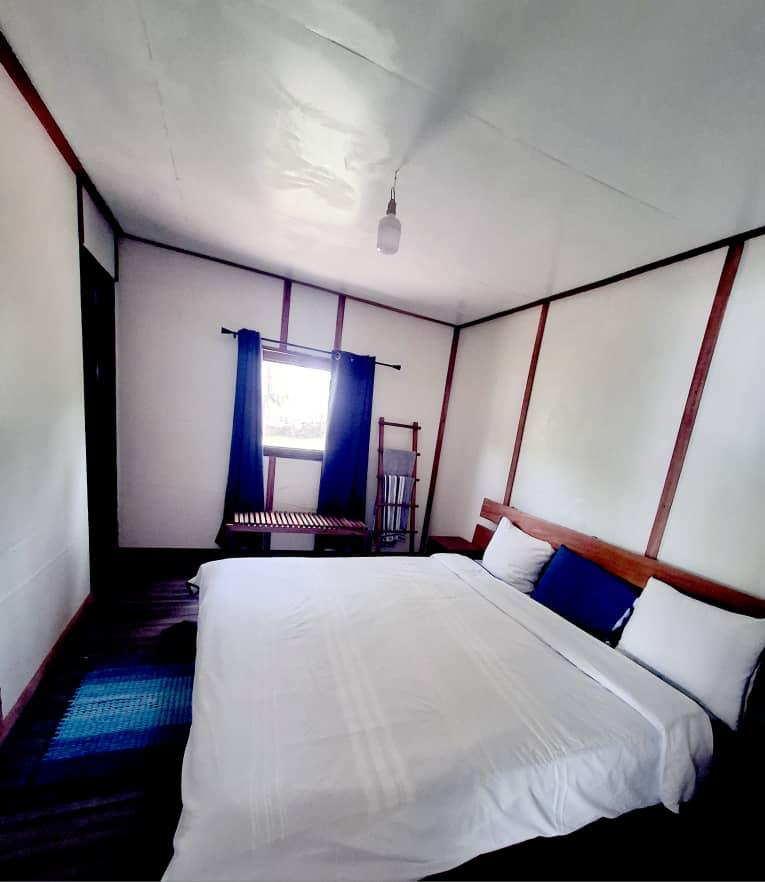 A spacious bedroom features a large white bed with blue and white pillows, a wooden headboard, and a window with blue curtains.
